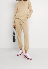 Beige huvtröja med avslappnad passform, i kombination med matchande joggers. Vita, rosa och svarta sneakers. Svart handväska med korta remmar.