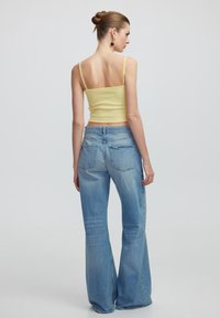 Top corto de punto amarillo con tirantes finos, combinado con unos pantalones vaqueros acampanados de color azul claro. Los pantalones cuentan con un diseño clásico de cinco bolsillos.