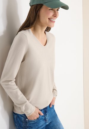 Donna che indossa un maglione a maniche lunghe color beige chiaro, un berretto verde e jeans blu, sorridente e in piedi contro un muro bianco con le mani nelle tasche.