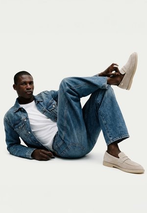 Man wearing light beige loafers, blue denim jacket and jeans, lying on floor with one leg bent and hand touching shoe.
