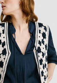 Kvačkana prsluk s cvjetnim uzorkom u krem i tamnoplavoj boji, nosi se preko tamnoplave bluze s naborima, koja ima volanasti ovratnik i detalj s mašnom.