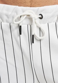Shorts de sport blancs avec des rayures noires verticales, une taille à cordon de serrage en corde blanche et des œillets noirs pour l'ajustement.