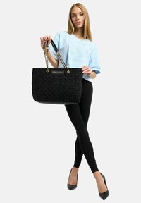 Schwarze gesteppte Tragetasche mit goldfarbenen Beschlägen und Kettenriemen, mit strukturierter Oberfläche und einem Markenlogo auf einer Plakette.