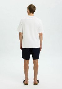 Biała koszulka z krótkim rękawem o luźnym kroju, zestawiona z granatowymi szortami. Materiał wydaje się gładki, z małym logo z tyłu.