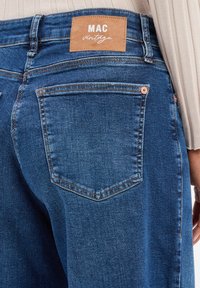 Jeans in denim blu scuro con vita alta, tasche posteriori con cuciture visibili e un'etichetta in pelle marrone con la scritta "MAC vintage."