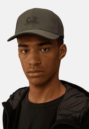 Giovane uomo che indossa un berretto C.P. Company grigio scuro, giacca nera e camicia nera, guarda dritto davanti a sé su uno sfondo semplice.