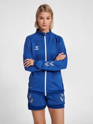 Junge Frau mit kurzen blonden Haaren, die eine blaue Sportjacke und passende Shorts mit weißen und silbernen Chevron-Mustern trägt.