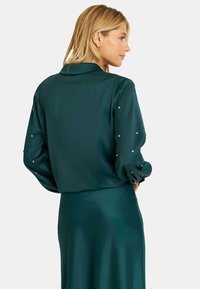 Tealte blouse met lange mouwen en een kraag, met parelachtige versieringen op de mouwen. Soepele, satijnen textuur en een comfortabele pasvorm.