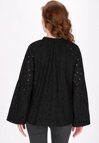 Blusa nera con maniche a campana lunghe, tessuto texturizzato con ricami e ritagli, scollatura arricciata e vestibilità leggermente ampia.