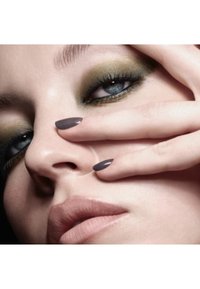 Gros plan sur un visage avec des ombres à paupières vert olive, des ongles gris et une couleur de lèvres naturelle, mettant en valeur une peau lisse et des traits bien définis.