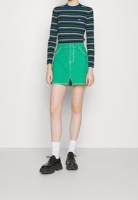 Jupe en denim vert avec une fente à l'avant, ourlet effiloché et poches découpées, associée à une chemise à manches longues bleue rayée et des chaussures plateformes noires.