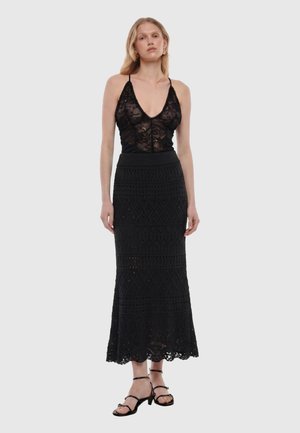 Donna che indossa un top nero in pizzo senza maniche con spalline sottili e una lunga gonna nera strutturata con tacchi aperti e cinturini.