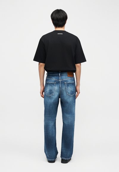 Άτομο που βλέπουμε από πίσω, φοράει μαύρο κοντομάνικο πουκάμισο και ξεθωριασμένο μπλε τζιν με ίσια πόδια, στέκεται σε απλό φωτεινό φόντο.