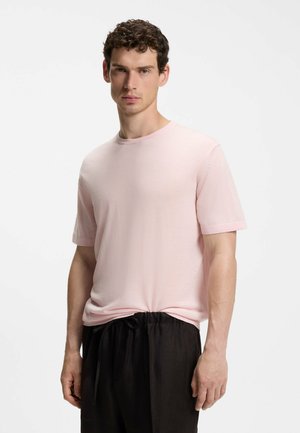 Mann mit kurzen, lockigen Haaren trägt ein hellrosa T-Shirt mit kurzen Ärmeln und weite schwarze Jogginghosen mit Kordelzug, stehend vor einem schlichten weißen Hintergrund.