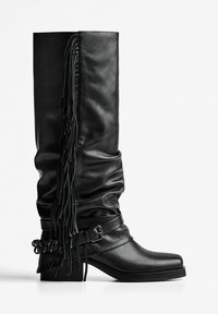 Black leather knee-high boot with slouchy design, fringe detail on the side, and a wide strap with a buckle. Chunky block heel.