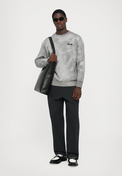 Mann in grauem MSGM-Sweatshirt, schwarzen Hosen, schwarz-weißen Loafers, Sonnenbrille und mit einer großen schwarzen Schultertasche vor weißem Hintergrund.