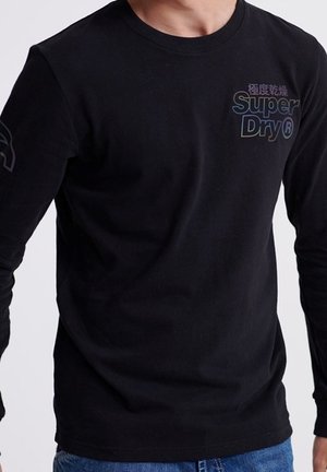 Mężczyzna w czarnej koszuli z długim rękawem z logo "Super Dry" z przodu, w połączeniu z niebieskimi dżinsami, stojący na jednolitym tle.