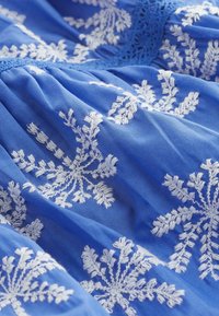 Blaue Stoff mit aufwendiger weißer Blumenstickerei, weicher Textur und dekorativem Spitzebesatz am Halsausschnitt.