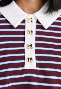 Bordo in svetlo modra prugasta polo majica z belim ovratnikom in štirimi zlatimi gumbi v obliki mašne po sprednjem zapenjanju.
