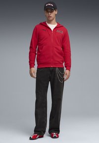 Κόκκινο φούτερ με φερμουάρ και λογότυπο στο στήθος, μαύρο τζιν με λεπτομέρεια αλυσίδας και κόκκινα αθλητικά παπούτσια. Μαύρο καπέλο με το κείμενο "AC Milan".