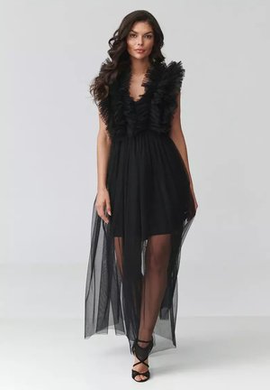 Kobieta o długich ciemnych włosach ubrana w czarną sukienkę bez rękawów z marszczonym biustem i przezroczystą, sięgającą do podłogi warstwą, czarne sandały na obcasie, stojąca na szarym tle.