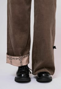 Barna kordbársony nadrág széles szárakkal, mintás rózsaszín szegéllyel. Fekete fűzős csizmával viselve, amely a texturált anyag részleteit mutatja.