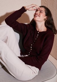 Cardigan en maille bourgogne avec des détails de boutons, porté sur un pantalon beige clair, devant un mur texturé. Texture en laine douce, coupe décontractée.