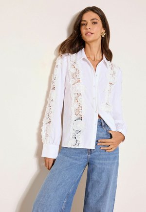Žena v bielej blúzke s dlhým rukávom s panelmi zo kvetín čipky vpredu a na rukávoch, v kombinácii s modrými vysokými džínsami.