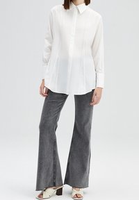 Witte langwerpige shirt met kraag, scherpe snit en knoopsluiting aan de voorkant. Gekoeld met uitlopende grijze jeans en lichtgekleurde gespierd muiltjes.