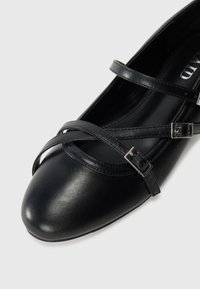 Scarpa piatta nera in pelle con due sottili cinturini regolabili e fibbie argentate sulla punta e sul collo del piede su uno sfondo bianco.