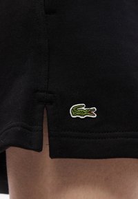 Μαύρο τελείωμα ενδύματος με μικρό πράσινο κεντημένο λογότυπο κροκοδείλου κοντά σε πλαϊνή σχισμή, που ακουμπά πάνω σε γυμνό δέρμα.