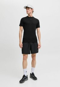 Schwarzes, kurzärmliges Sportshirt mit einem Logo auf der Brust, kombiniert mit schwarzen Shorts. Das Modell trägt eine graue Mütze und schwarze Sportschuhe.