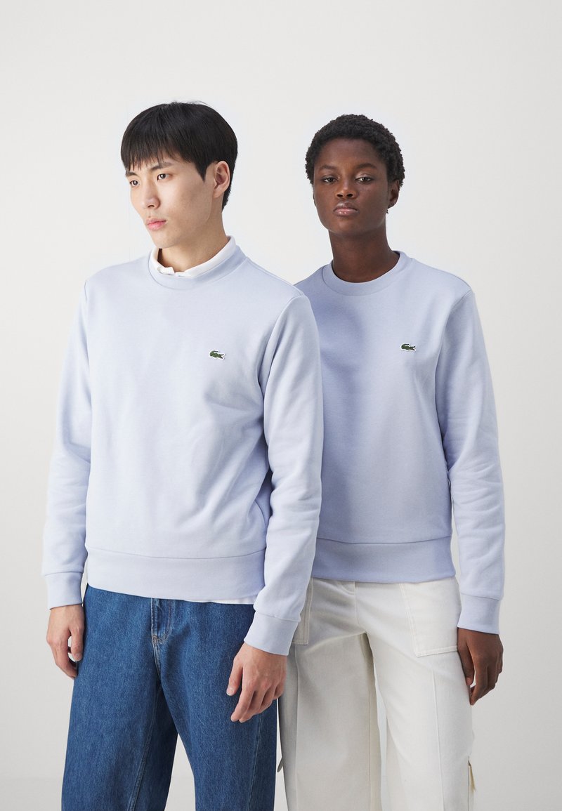 Lichtblauwe sweaters met ronde hals en groene geborduurde logo's. Het ene model draagt jeans, het andere model draagt crèmekleurige broeken.