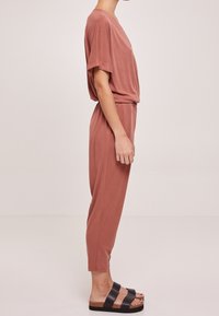 Combinaison couleur rouille avec manches courtes, taille élastique et jambes fuselées. Portée avec des sandales noires à bretelles épaisses et semelle en liège.