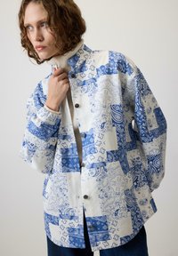 Jachetă din patchwork albastru și alb, cu modele paisley, butoni tip snap și o croială lejeră. Țesătura are o suprafață texturată, cu cusături vizibile.