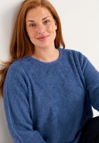 Blauer flauschiger Pullover mit Rundhalsausschnitt und langen Ärmeln, der eine weiche Textur und eine lockere Passform aufweist. Getragen mit dunklen Hosen.