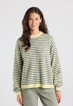 Vrouw met lang donker haar die een geel-grijs gestreepte trui en lichte broek draagt, staand tegen een effen achtergrond.