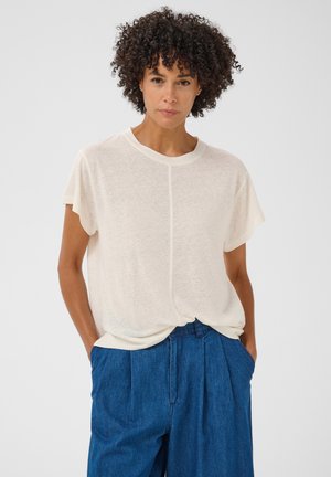 Femme aux cheveux bouclés portant une chemise blanche ample à manches courtes et un pantalon bleu taille haute, debout les mains dans les poches.