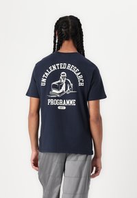 Navy blue cotton t-shirt featuring a graphic design of a person at a computer, with "UNTALENTED RESEARCH PROGRAMME" text on the back.