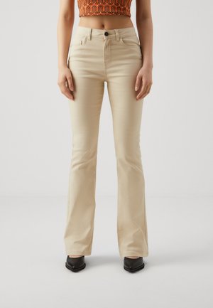 Femme portant un pantalon flare beige taille haute, un haut court à motif orange et des chaussures noires à bout pointu, debout devant un fond uni.