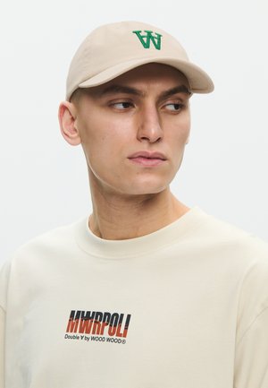 Ung mand iført en beige kasket med grønt "W" logo og en cremefarvet sweatshirt med teksten "MWRPOLI Double W by WOOD WOOD".