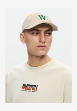 Ung mand iført en beige kasket med grønt "W" logo og en cremefarvet sweatshirt med teksten "MWRPOLI Double W by WOOD WOOD".