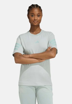 Mujer con el cabello trenzado que lleva una camiseta y pantalones Adidas de color gris claro, con los brazos cruzados, mirando hacia adelante con expresión neutral.