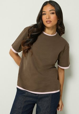 Femme aux longs cheveux foncés portant un t-shirt marron avec des bordures rose clair et un jean bleu foncé, posant devant un fond clair uni.