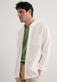 Vit skjorta med knäppning över en grön t-shirt. Skjortan har en enda bröstficka och är gjord av ett mjukt, slätt tyg. Beiga byxor fulländar outfiten.