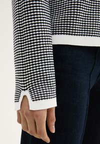 Gestrickter Pullover in Schwarz und Weiß mit kariertem Muster. Mit einem weißen, gerippten Saum und einem Ausschnitt am Ärmel.