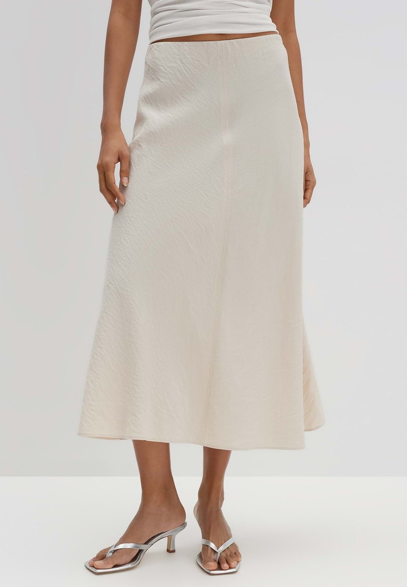 Jupe midi couleur crème en tissu texturé, avec une taille ajustée et un ourlet évasé, accompagnée de sandales à talons métalliques.