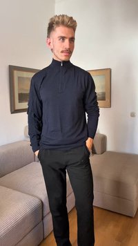 Mornarsko modra zip-up jopica z dolgimi rokavi, teksturirana tkanina, v kombinaciji s črnimi hlačami. Ozadje vsebuje svetlo obarvano pohištvo in umetniška dela.