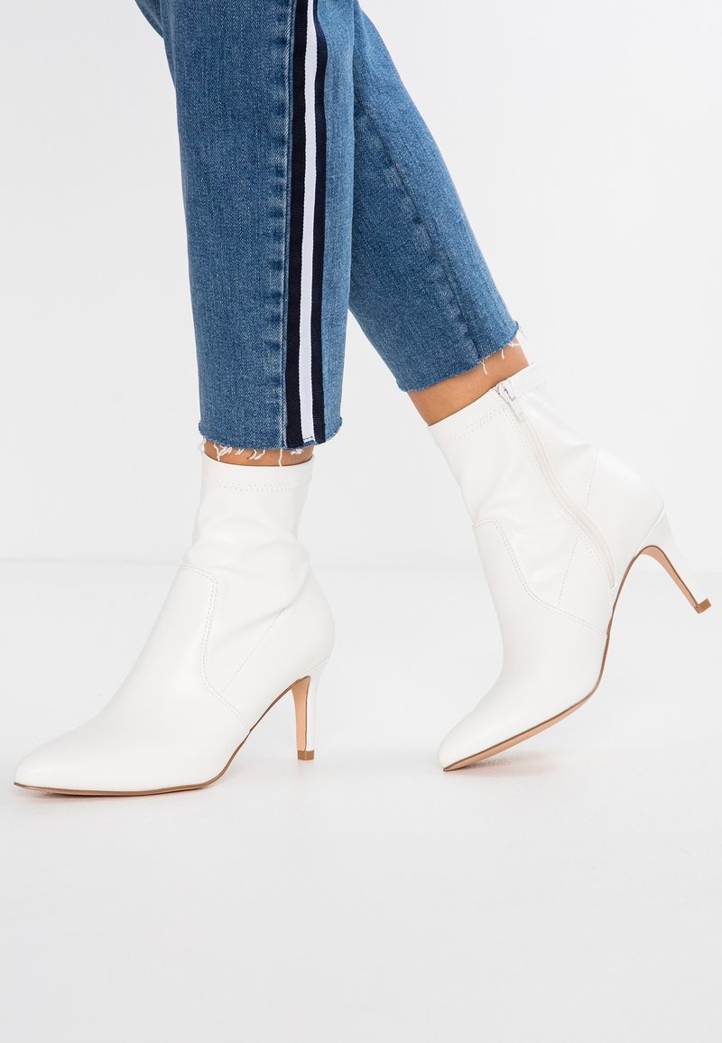 Bottines blanches en cuir mi-mollet avec des bouts pointus et des talons fins, portées avec un jean muni d'une bande latérale noire et blanche.