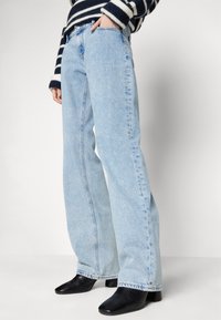 Persona que lleva jeans de tiro alto y pierna ancha en color azul claro, botas de tobillo negras con puntera cuadrada, y un suéter a rayas en blanco y negro con una mano en el bolsillo.